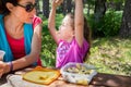 Funny girl playing with macaroni in her mouth looking at her mother eating from a plastic lunchbox in a table picnic in country