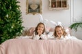 Funny giggling girls are lying on bed by Christmas tree and looking at camera