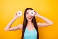 Funny girl in blue singlet with beaming smile holding white tasty donuts near eyes, fooling around on bright yellow background Royalty Free Stock Photo