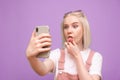 Funny girl with light hair doing a selfie on a purple background and making a funny face. A funny teen girl takes a selfie on a