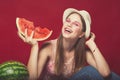 Funny girl with pink make up, wearing jeans, hat and top, posing at red studio background, holding slice watermelon, looking at Royalty Free Stock Photo