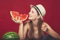 Funny girl with pink make up, wearing jeans, hat and top, posing at red studio background, holding slice watermelon, looking on Royalty Free Stock Photo