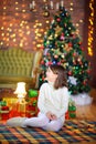 A funny girl sits on the floor next to gifts on the background of bright holiday lights