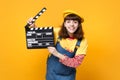 Funny girl teenager in french beret, denim sundress holding classic black film making clapperboard isolated on yellow Royalty Free Stock Photo