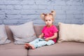 A funny girl of two years from birth with white hair and in a red shirt and blue pants plays a mobile phone on the couch in a room Royalty Free Stock Photo