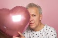 funny gray-haired elderly pensioner man holding a pink heart-shaped balloon in his hands on a pink background Royalty Free Stock Photo