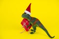 Green dinosaur toy in little santa claus hat with red gift box on vibrant yellow background