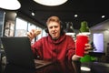 Funny guy in headphones and a burger with a drink in his hands, sitting in a cafe at the table near the laptop Royalty Free Stock Photo