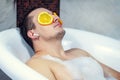 Funny guy washing his bath. Listens to music with wireless headphones. Relaxed from spa procedures on face with orange round slice Royalty Free Stock Photo