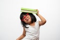 Funny and Happy Asian little preschool girl wearing red glasses holding a green book on the head, on white isolated background. Royalty Free Stock Photo