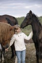 Funny happy child in a white sweater and jeans standing among horses foals on the farm smiling. Lifestyle portrait Royalty Free Stock Photo