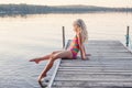 Funny happy cute Caucasian blonde girl child sitting on wooden dock pier by lake. Pensive kid in swimsuit splashing with legs in Royalty Free Stock Photo