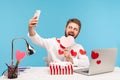 Funny happy man with beard in white shirt with red sticky hearts making selfie with pink heart and striped giftbox sitting at Royalty Free Stock Photo