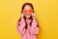 Funny happy smiling little girl wearing pink sweatshirt covering her eyes with red little hearts isolated over yellow background Royalty Free Stock Photo