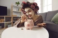 Smiling young woman with face mask and hair curlers putting coins in her piggy bank Royalty Free Stock Photo