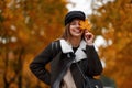 Funny happy young woman in stylish outerwear in a vintage black hat posing in a park. Cheerful girl model holds an orange maple Royalty Free Stock Photo