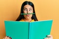 Funny hispanic woman reading a book doing crazy gesture with pencil over mouth like mustache, playful and positive on free time,