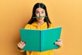 Funny hispanic woman reading a book doing crazy gesture with pencil over mouth like mustache, playful and positive on free time,