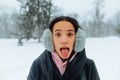 Funny Hispanic woman in warm clothes looks at the camera and makes a funny face showing her tongue on the background of a blizzard