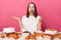 Funny hungry brown haired woman in white t shirt sitting at table with sweets isolated over pink background posing with mouth full