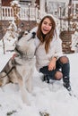 Funny husky resting after game in snowy park. Outdoor portrait of elegant white woman in ripped jeans sitting on the