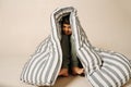 Funny indian boy sitting under a striped mattress over beige background.