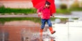 Funny kid girl in a red jacket and rubber boots with an umbrella walks fun through the puddles Royalty Free Stock Photo