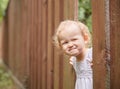 Funny kid in light clothes looks out of a hole in the fence. Summer sunny day. Royalty Free Stock Photo