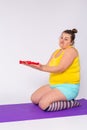 Funny large woman with doubt expression holding dumbbells, sitting on violet fitness mat, gray background. Plus size