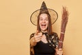 Funny laughing young woman wizard wearing witch costume holding in hand broom  over beige background pointing to camera Royalty Free Stock Photo