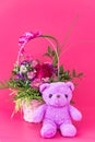 Funny lilac teddy bear with basket of flowers on pink background. Cute greeting postcard