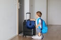 Funny little baby boy in fathers shoes with big backpack, suitcase and spoon in his hand staying in hallway looking at door ready