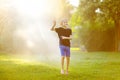 Funny little boy playing with garden sprinkler in sunny city park. Elementary school child laughing, jumping and having fun with