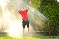 Funny little boy playing with garden sprinkler in sunny city park. Elementary school child laughing, jumping and having fun with