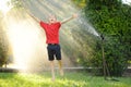 Funny little boy playing with garden sprinkler in sunny city park. Elementary school child laughing, jumping and having fun with