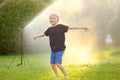Funny little boy playing with garden sprinkler in sunny city park. Elementary school child laughing, jumping and having fun with