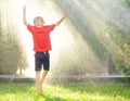 Funny little boy playing during a rain in sunny city park. Elementary school child laughing, jumping and having fun with spray of