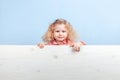 Funny little curly girl in a striped red and white dress and blue flower on her hair stands behind the wooden board Royalty Free Stock Photo