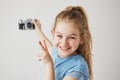 Funny little girl with blue eyes and light hair smiles, holding photocamera in her hand, showing v-sign, going to take