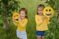 Funny little girl girlfriends hold paper faces of emoji and laugh merrily standing under a tree in the park.