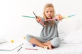 Funny little girl in gray pajamas playing with colorful pencils, looking at camera, sitting on bed Royalty Free Stock Photo