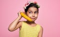 A funny little girl holds a banana, like a phone, isolated on pink background. A happy kid in a yellow dress pretends to talk on a Royalty Free Stock Photo