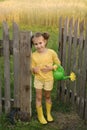 A funny little girl with a plastic watering can stands at the gate of an old wooden fence leading to the garden Royalty Free Stock Photo