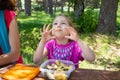 Funny little girl playing with macaroni eating from a plastic lunchbox next to her mother in a table picnic in countryside Royalty Free Stock Photo