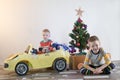 Funny little smiling kids driving toy car with Christmas tree. Happy child in colour fashion clothes bringing hewed xmas tree from Royalty Free Stock Photo