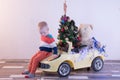 Funny little smiling kids driving toy car with Christmas tree. Happy child in colour fashion clothes bringing hewed xmas tree from Royalty Free Stock Photo