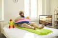Funny man sitting on bed with inflatable swim ring, sipping juice and watching travel TV show