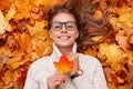 Funny model young woman with a cute smile in a glasses in a sweater with a maple leaf in hands lies on golden foliage. Pretty