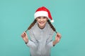 Funny optimistic teenager girl in Santa Claus hat, looking at camera and pulling her pigtails aside. Royalty Free Stock Photo