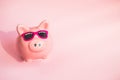Funny piggy Bank in sunglasses on pink background, sunlight, copy space, money saving concept for summer vacation